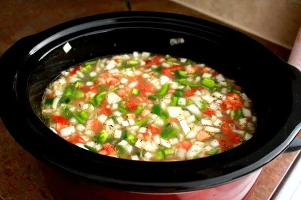 Overhead flat-lay photograph of a black slow cooker filled with diced tomatoes, onions, and green peppers in broth on a warm-toned countertop, dominated by deep reds, fresh greens, and creamy whites, conveying a wholesome home-cooked mood.